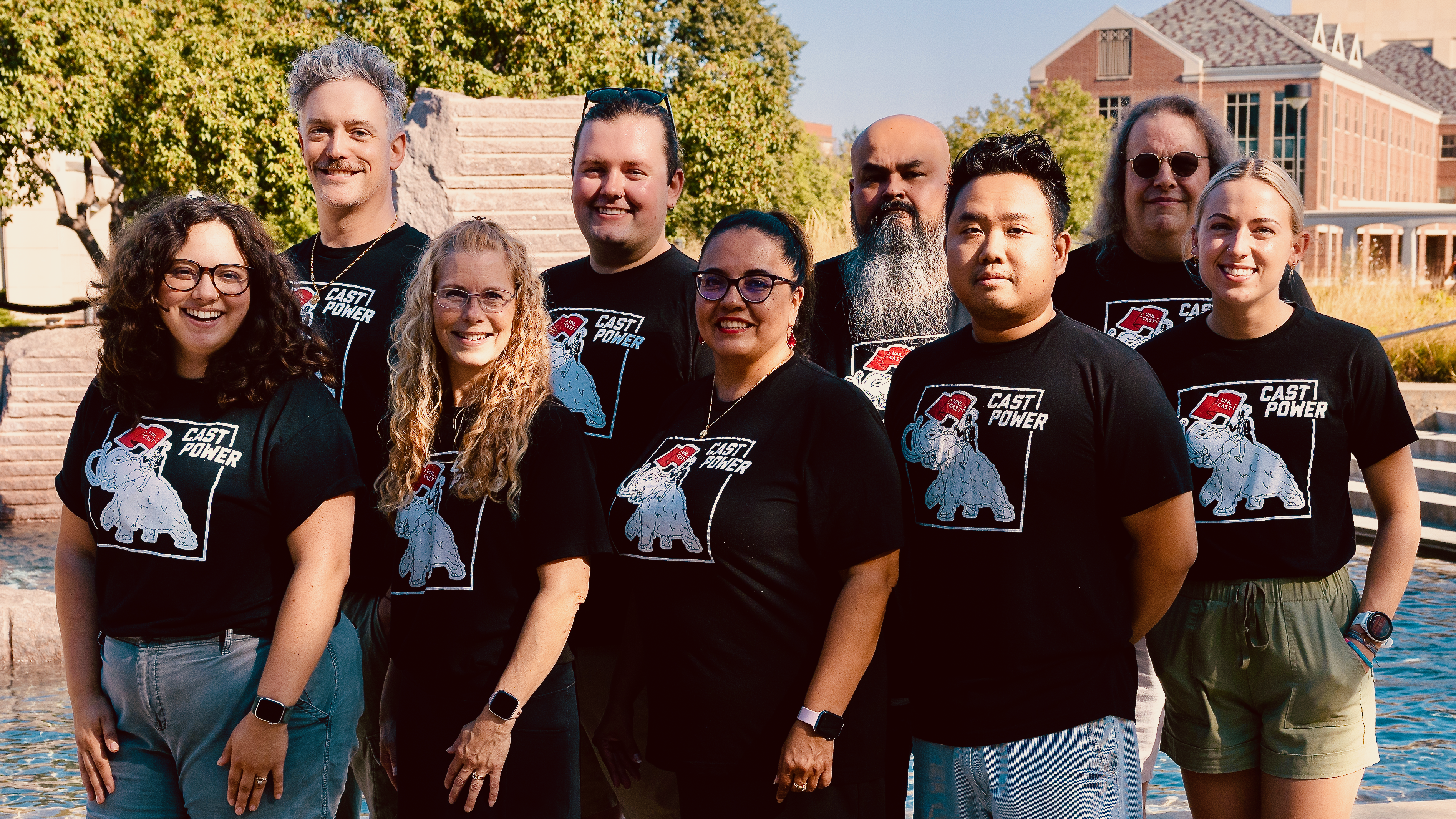 A group of nine CAST staff members stand together outside by the Nebraska Union fountain. They are smiling and wearing matching black CAST POWER t-shirts with a mammoth logo.