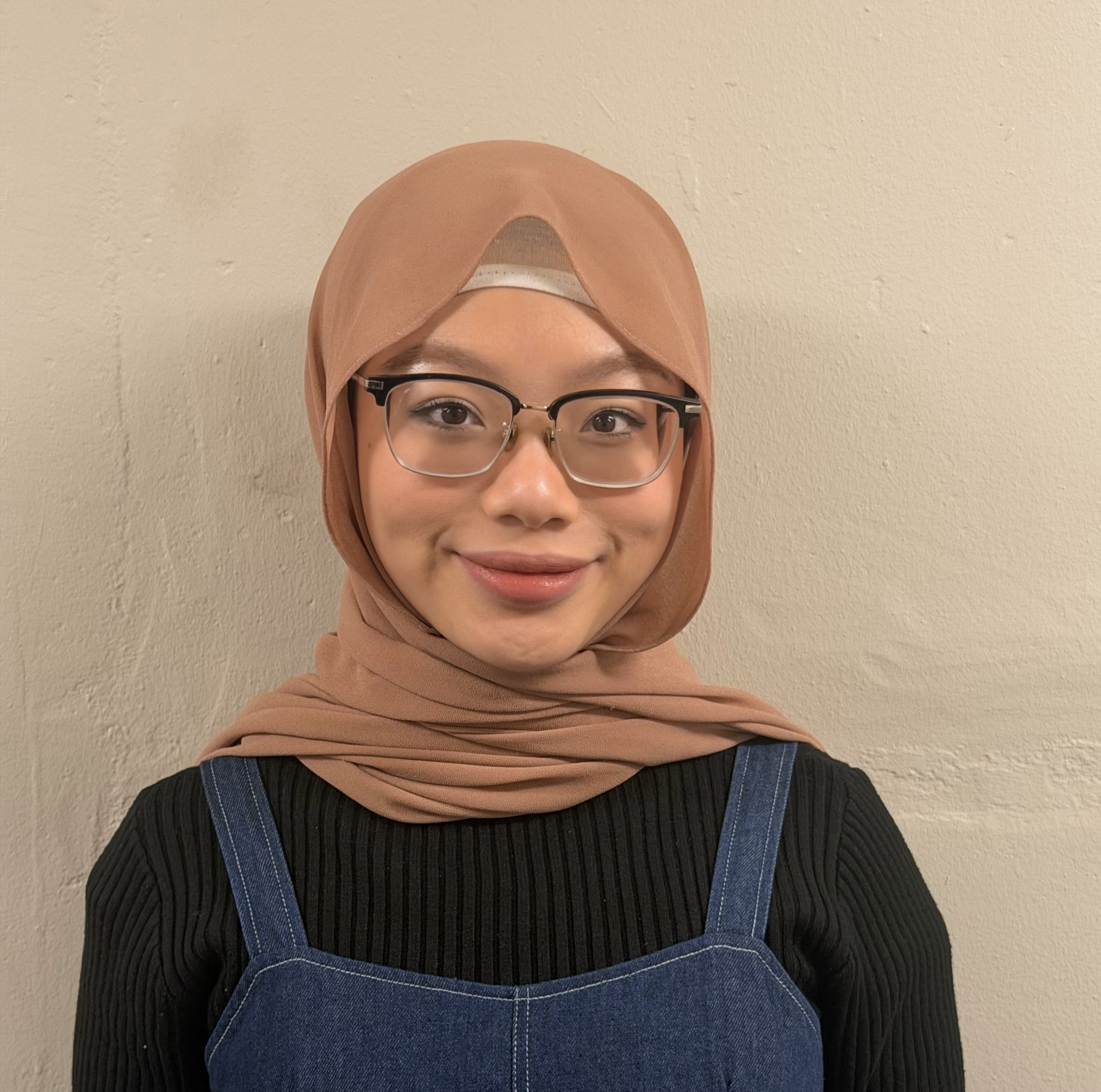 Woman wearing a light brown hijab, glasses, and a black top with a denim dress, smiling warmly.