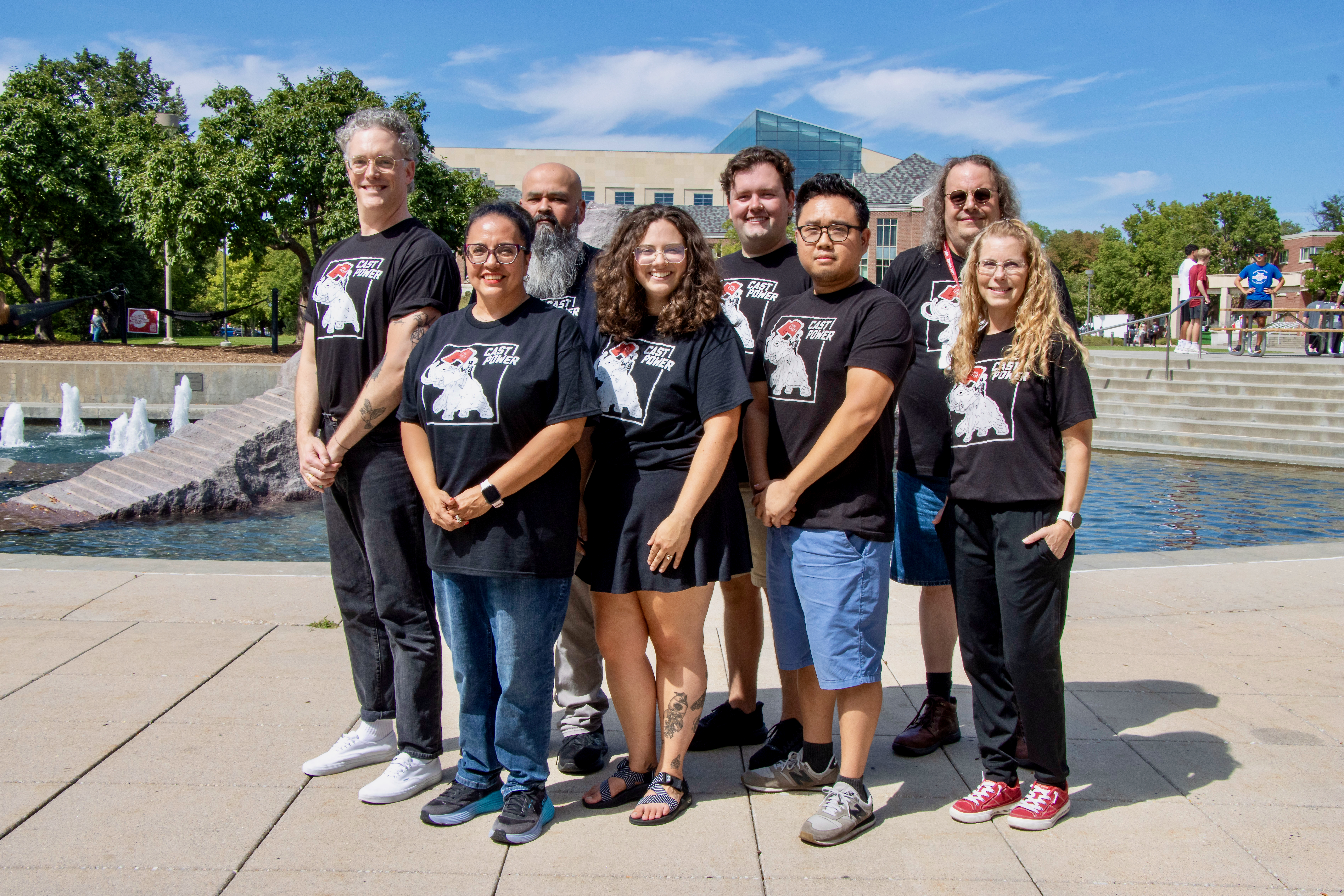 CAST staff pose together near Broyhill Fountain on the University of Nebraska–Lincoln campus, with Howard L. Hawks Hall in the background. Front row, from left: Mari Beck, Abby Roskos, Donny Bui, and Lynnette Sallinger. Back row, from left: Rob Russell, Joel Cardenas, Jordan Heim, and Dan Hutt. Everyone is wearing black CAST POWER shirts.