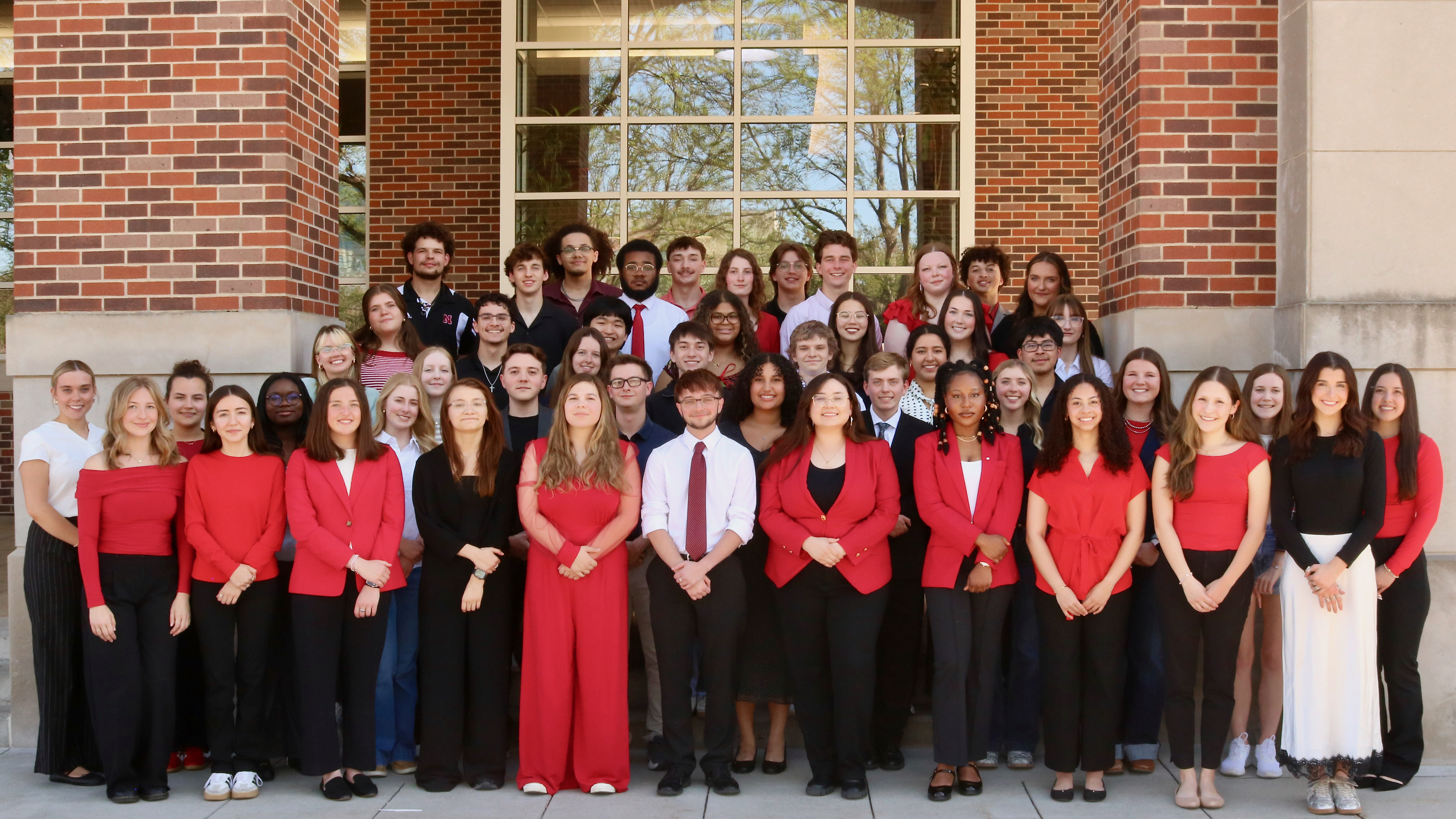 Large group of about 50 college students posed in multiple rows outside a brick campus building with tall windows. Most are dressed in red, black, and white outfits, with several in business casual attire such as blazers, dresses, and ties. The group stands on concrete steps, smiling and facing the camera, with trees reflected in the windows behind them.