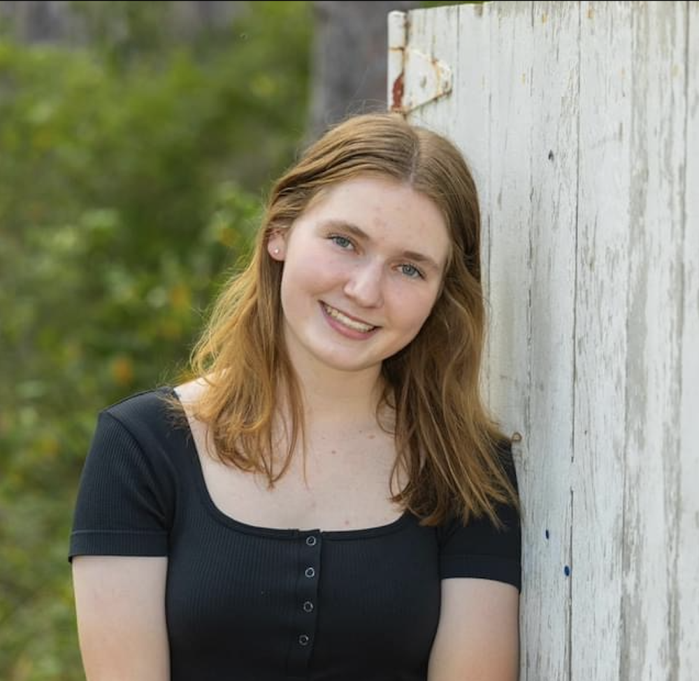 Smiling person leaning against a weathered white wooden wall with greenery in the background.