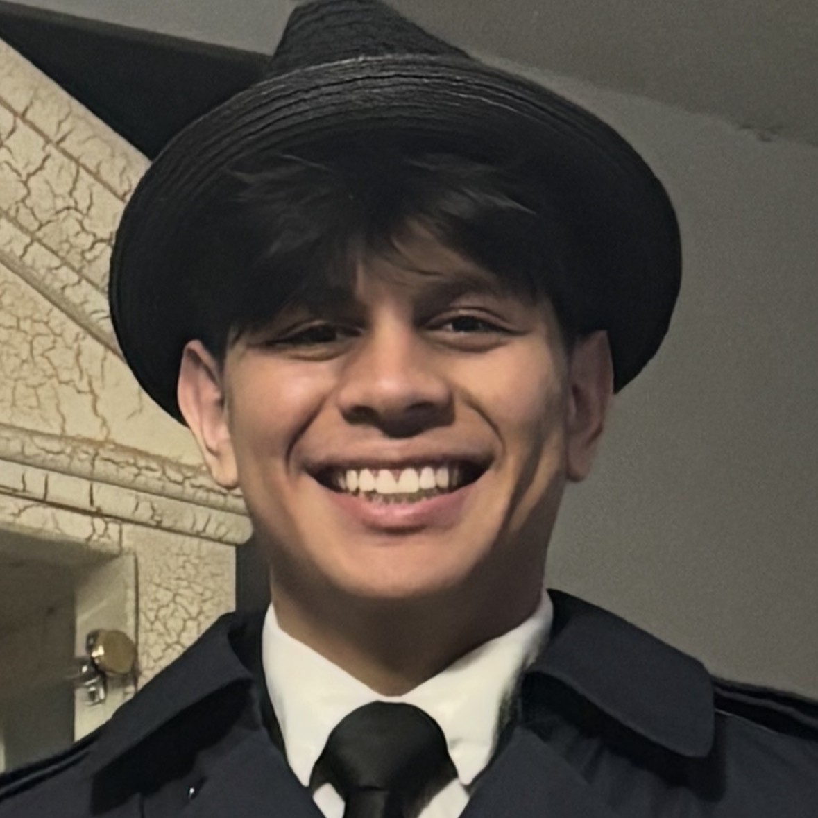 Smiling person wearing a black hat and formal attire indoors.
