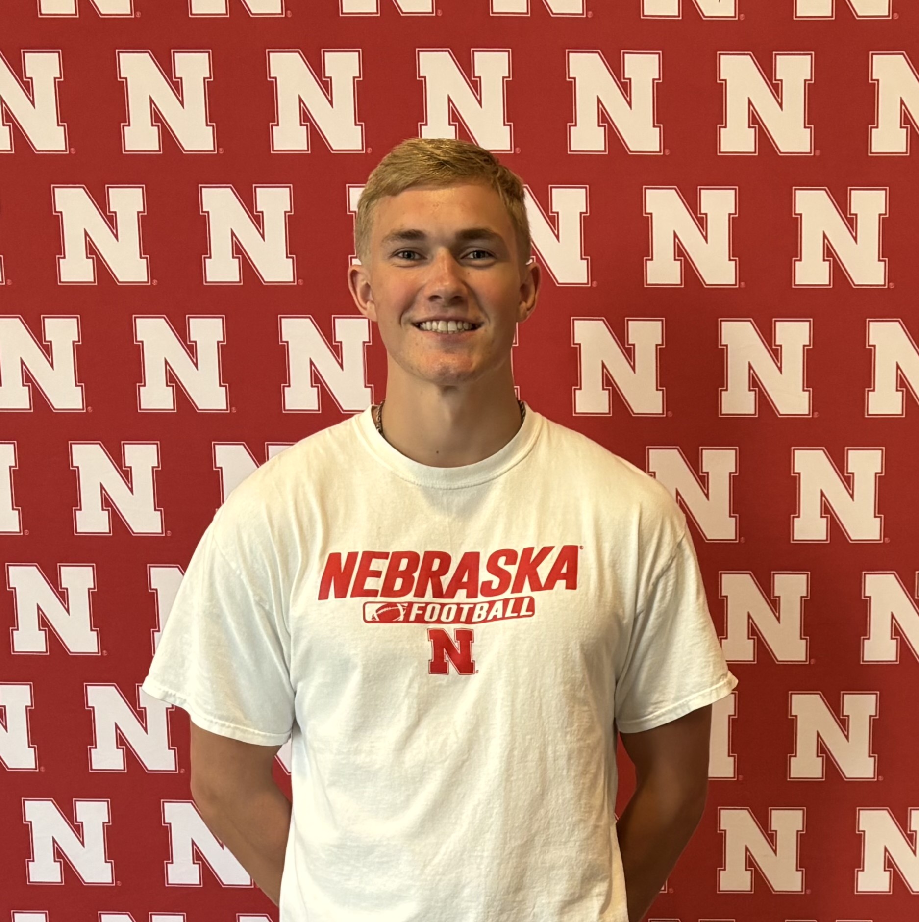 Man smiles in front of a red backdrop with repeating white "N" logos, wearing a Nebraska Football shirt.
