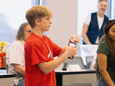 A student in a red shirt holds up a phone in a classroom, with other people in the background.