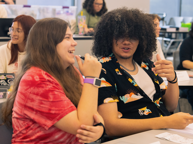 Two students sit at a classroom table talking and laughing. One wears a red tie-dye shirt and holds a pen, while the other wears glasses and a patterned black shirt. Other people are visible in the background.