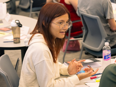 A student in glasses and a white hoodie speaks while gesturing with her hands at a classroom table, with notebooks, markers, and a water bottle in front of her.