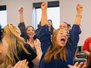 A group of students cheer indoors, with one woman in a blue sweater and glasses raising both arms high in celebration while smiling widely. Others around her clap and cheer with excitement.