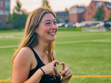 A young woman with long hair smiles while making a hand gesture in the shape of a heart on a sports field.