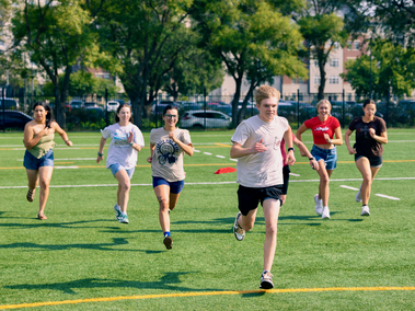 A group of students run across a sports field, with one student in a light shirt and black shorts leading ahead of the others. Trees, buildings, and parked cars are visible in the background.