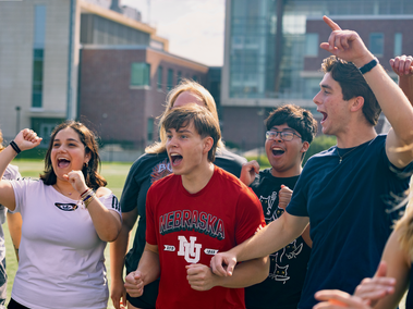 A group of students cheer outdoors on a sports field. A student in a red Nebraska shirt stands in the center with others around him smiling, clapping, and raising their arms in excitement.