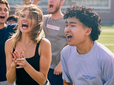 A group of students cheer energetically on a sports field. A young woman in a black tank top claps while shouting, and a young man in a light blue shirt yells alongside her, with others cheering in the background.