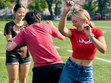 Three students laugh and dance on a sports field. A woman in a red “Nebraska” shirt smiles with her arms raised, while another student in a red shirt faces her, and a third student in glasses watches and smiles in the background.