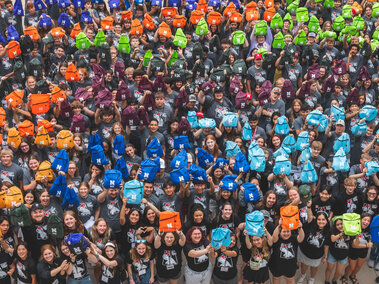 Large group of students standing together in matching shirts holding up different color drawstring bags.